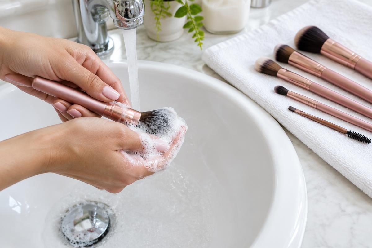 Hands washing a makeup brush over a porcelain sink with clean brushes on a towel